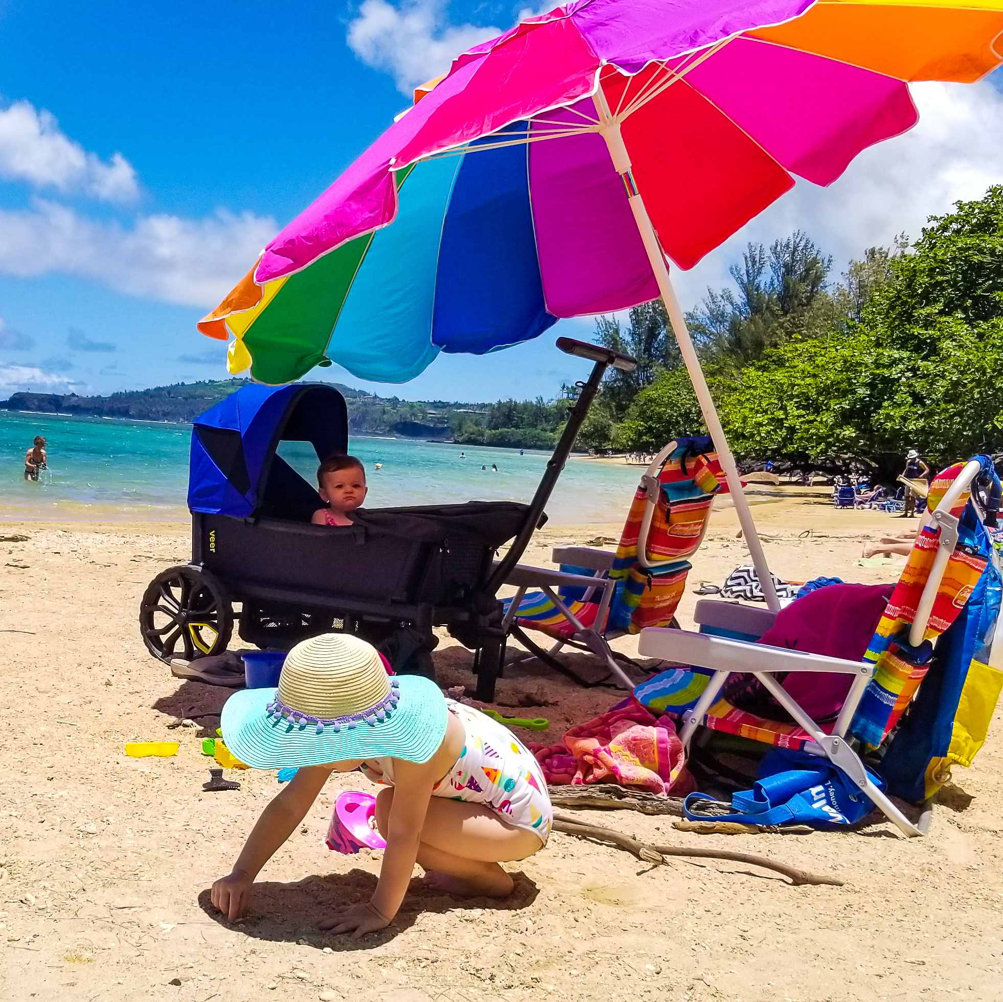brightly colored beach set up with umbrella, chairs, veer cruiser, toddler, and baby