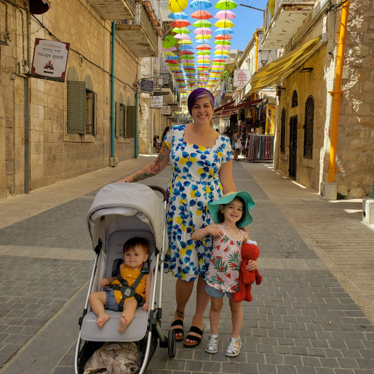 woman standing with baby in uppababy minu stroller and toddler holding elmo doll in front of street of umbrellas