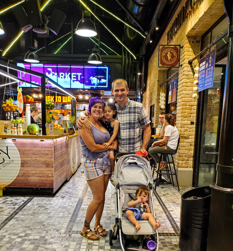 family smiling in front of food stalls and sarona market sign