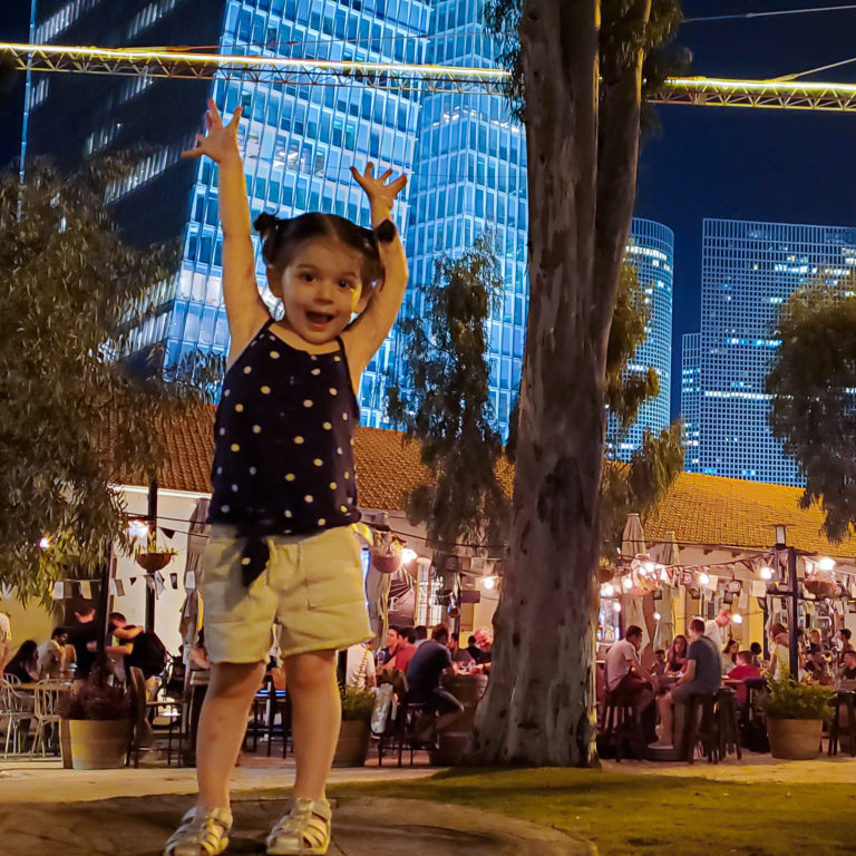 toddler waving arms in front of restaurant with skyscrapers in background