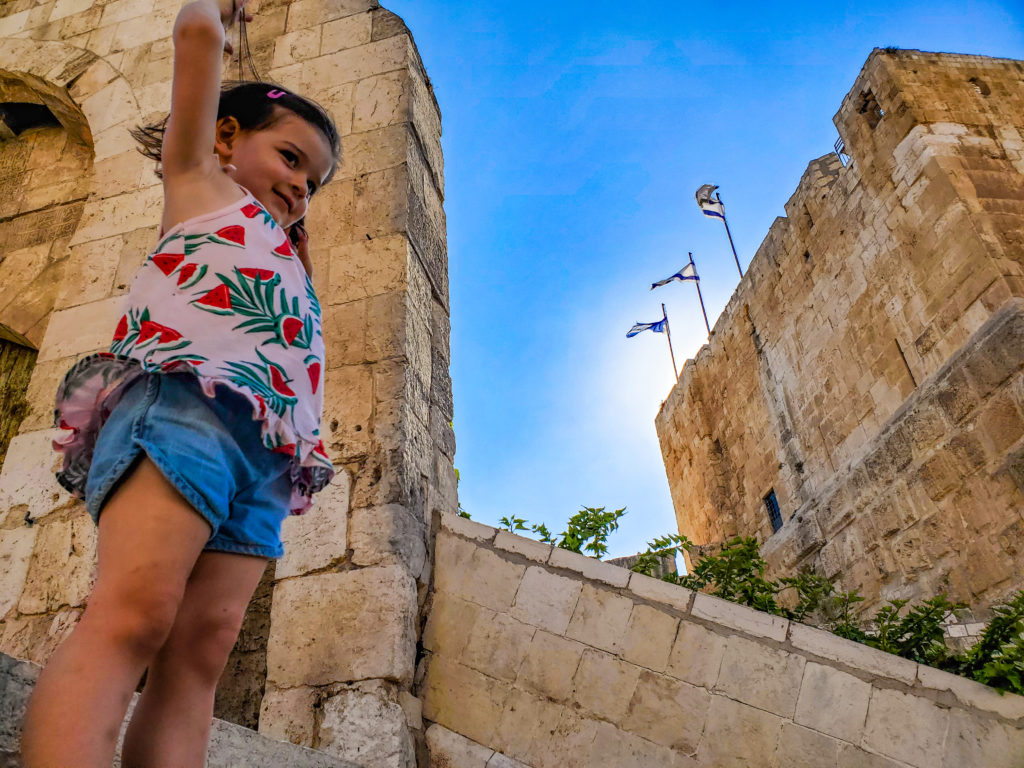 shot of toddler in front of ancient tower with three israeli flags flying from it