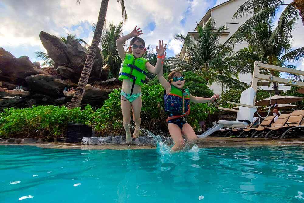 3 and 5 year old girls mid-jump into a pool, both wearing vests, with palm trees in background