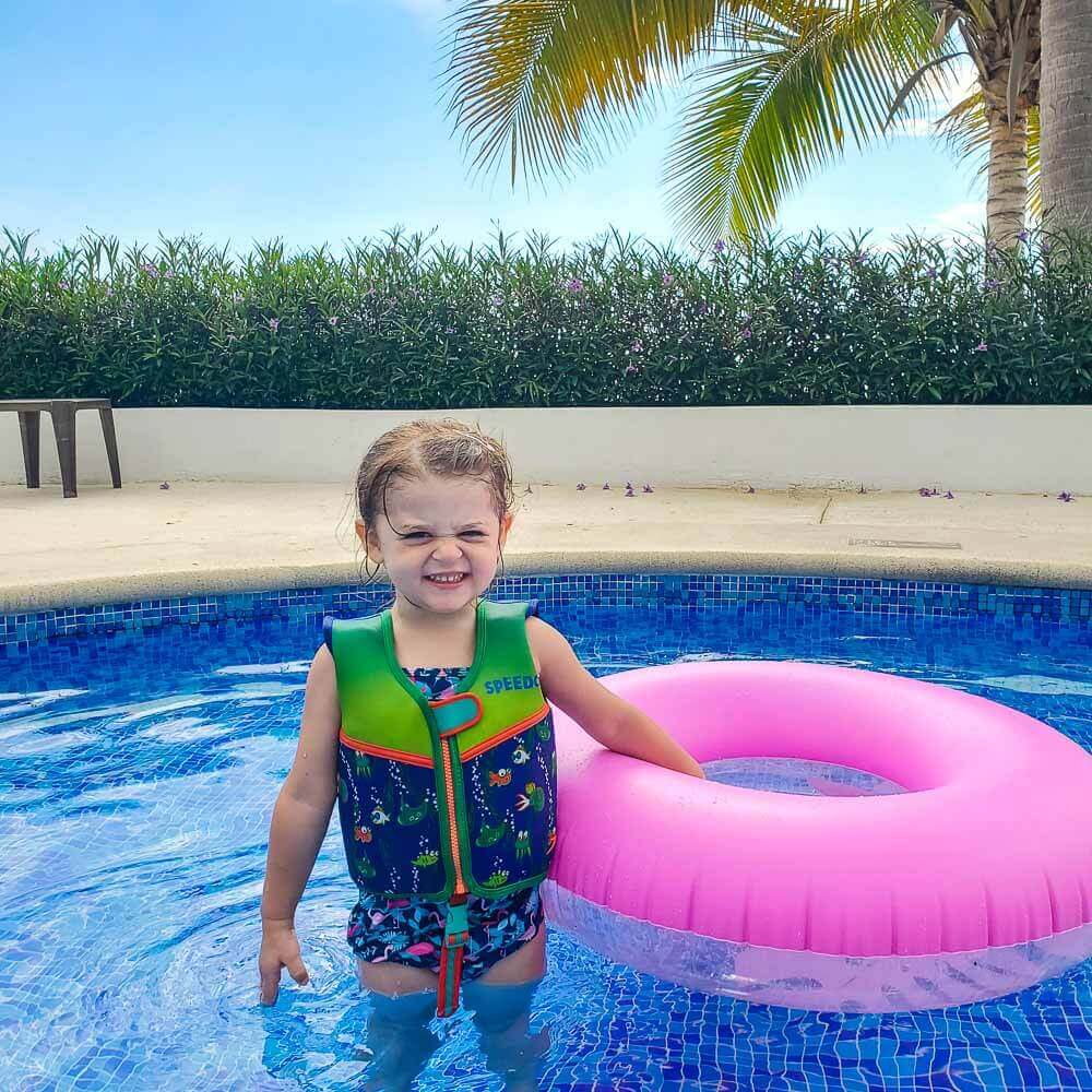 smiling toddler girl in shallow pool with pink floatie wearing a green and blue toddler swimming vest